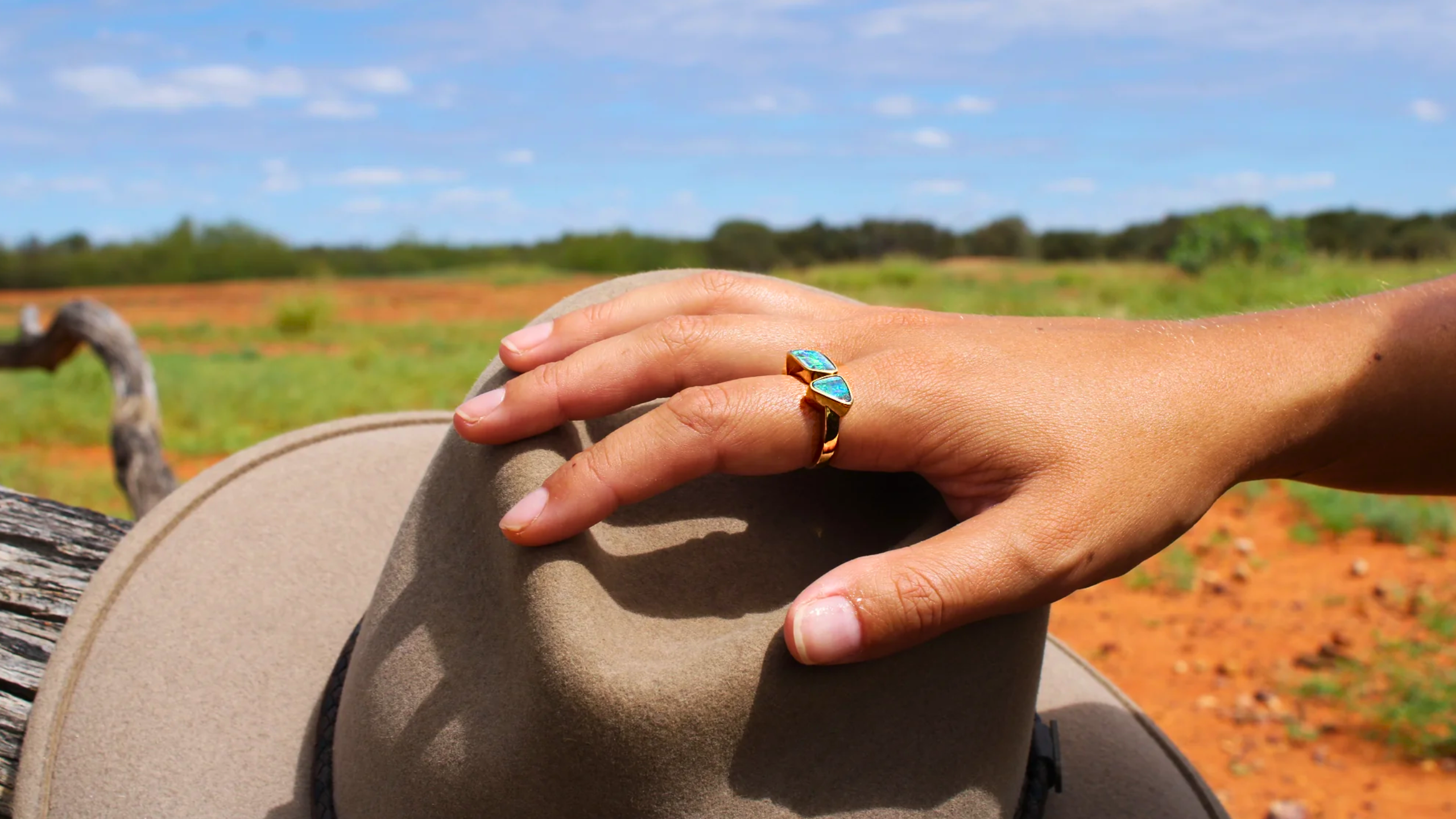 Hand wearing a gold ring with a turquoise stone on a beige hat against a rural landscape.