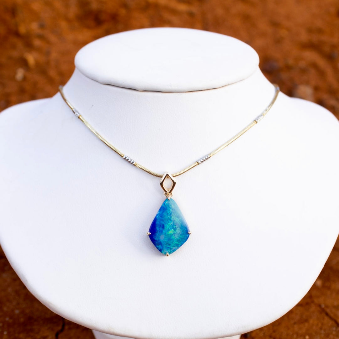 Pale sky blue to ocean blue Australian Boulder Opal cut in a diamond shape with a four claw setting and an open diamond shaped bale hanging above the stone. Set in 18K solid yellow gold. Some shimmering green colours flash across the face of the blue opal. The opal's colouring and patterns are reminscent of a shimmering aqua blue water reflecting in sunlight.