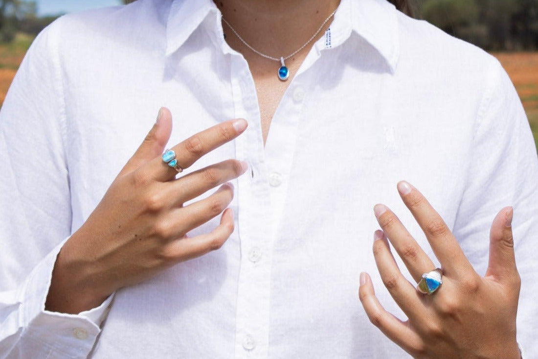 Person wearing a white shirt with a blue opal necklace and blue opal rings on her fingers, outdoors in the beautiful red dirt Australian outback landscape.