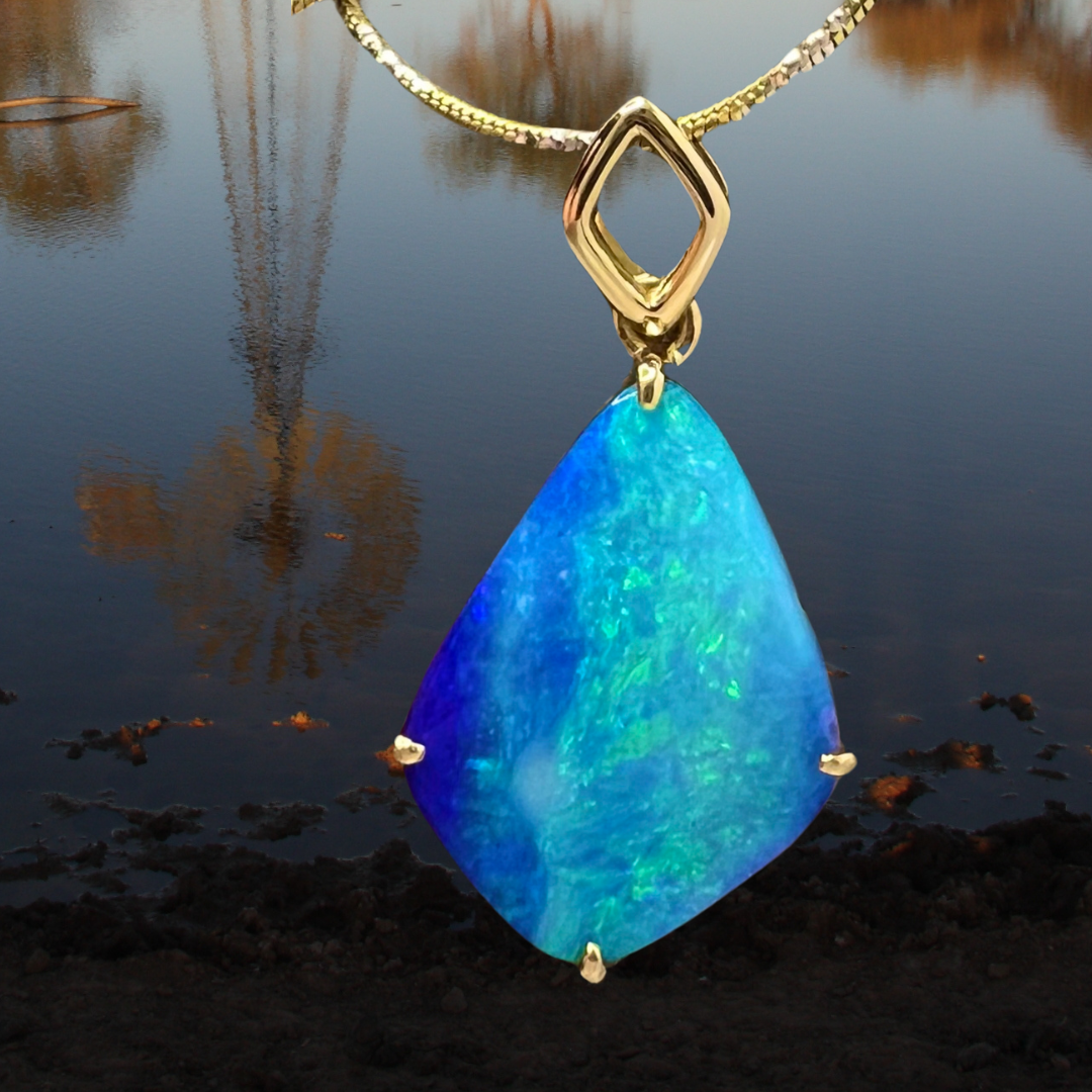 A bright blue with pale shimmering greens and deeper contrasting blue coloured opal hangs in front of a watery outback setting. The water reflection shows a wind mill in the natural scenery behind the necklacke. 