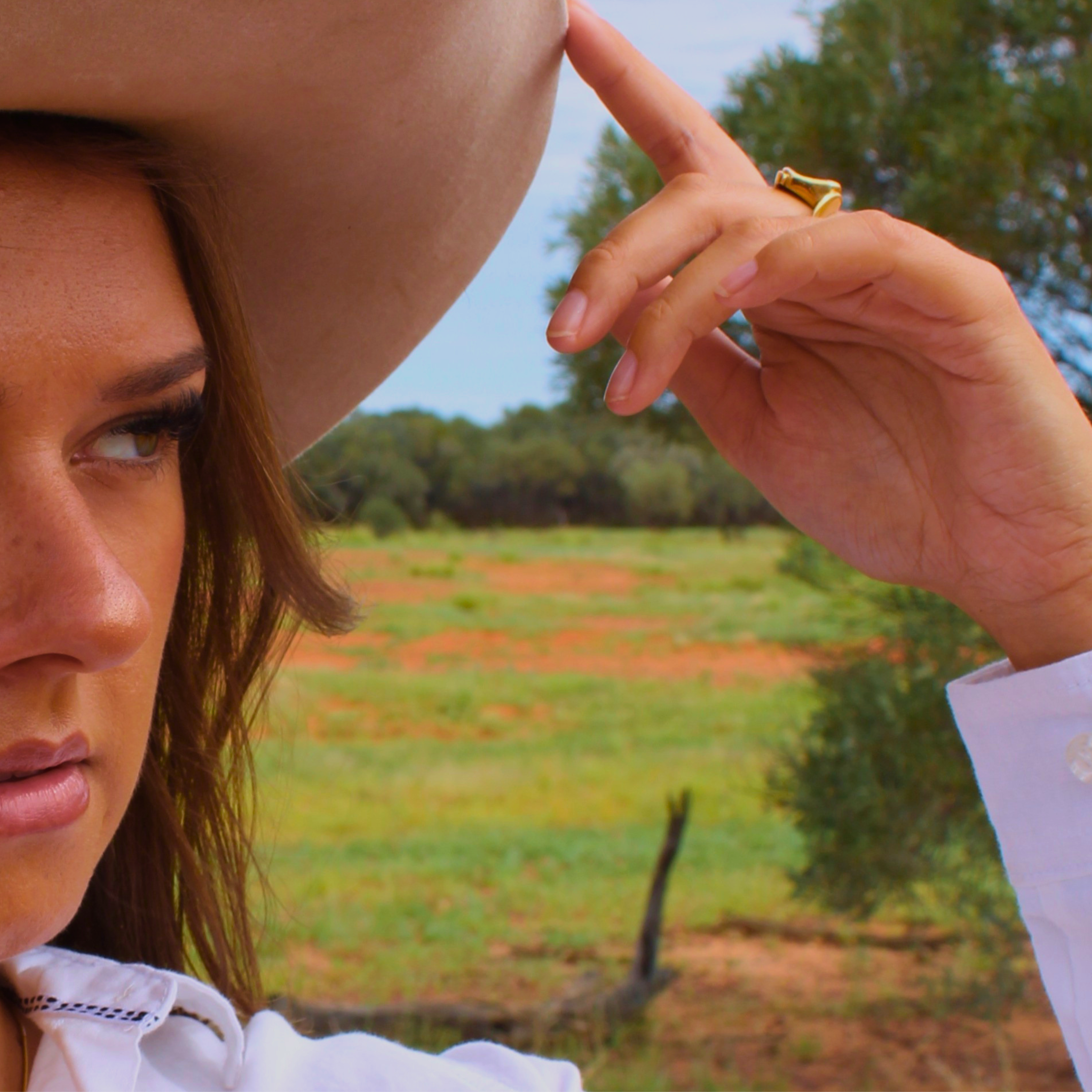 Woman wearing a cowboy hat and a gold ring with a scenic background of trees and open field.